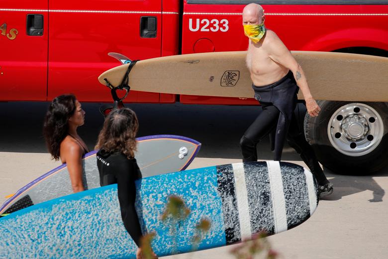   Một người lướt sóng lớn tuổi đeo khẩu trang đi ngang qua những người lướt sóng trẻ hơn không đeo khẩu trang ở bãi biển Encinitas, California. Ảnh: Reuters.  