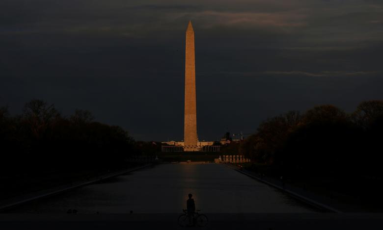   Một người đi xe đạp dừng trước Hồ bơi phản chiếu Lincoln Tưởng niệm, với Đài tưởng niệm Washington được thắp sáng bởi mặt trời lặn ở phía sau, tại Washington.  