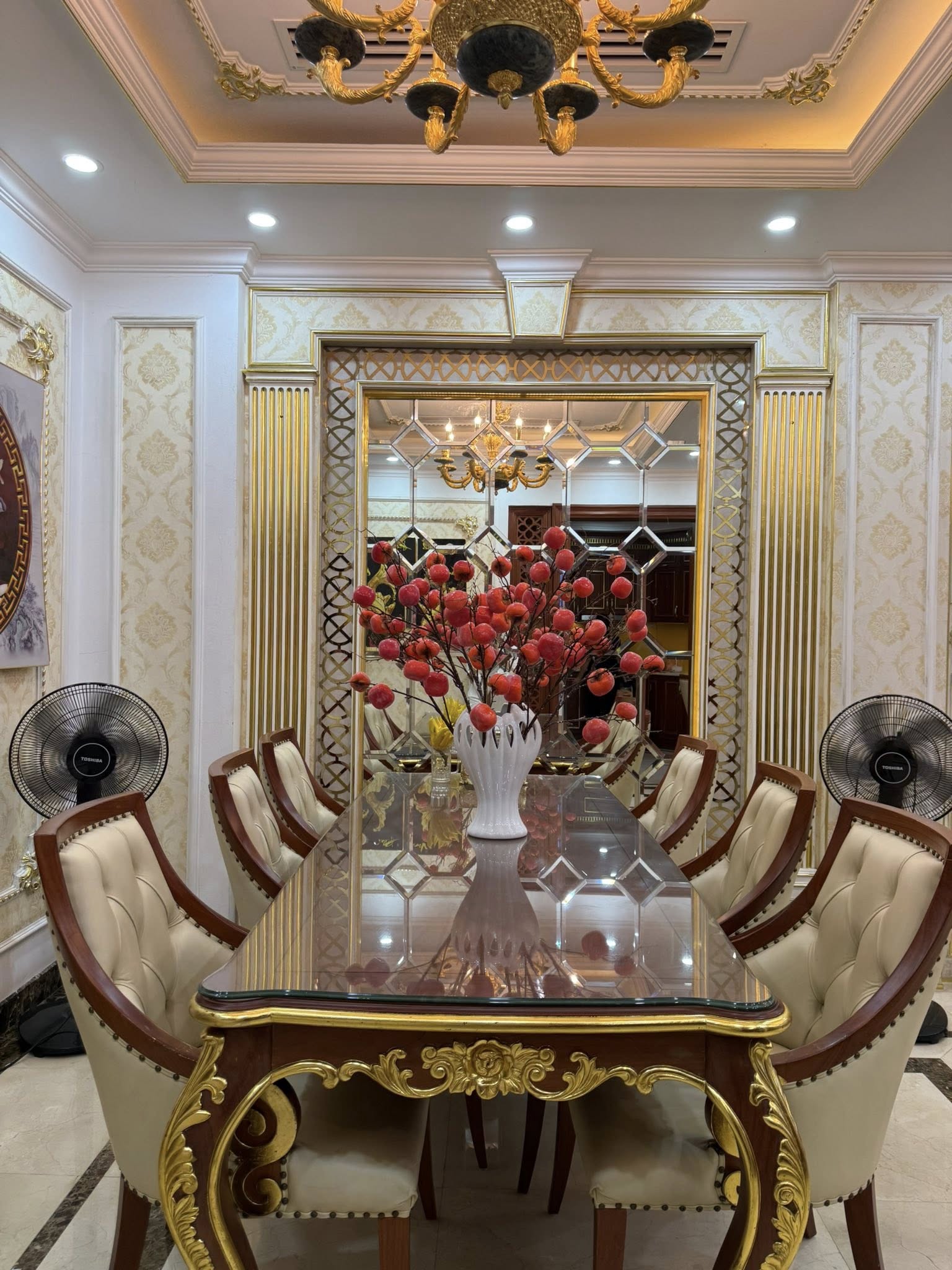 Khu vực bàn ăn tiếp tục sử dụng gương trang trí cỡ lớn, tạo hiệu ứng mở rộng không gian và tăng chiều sâu cho tổng thể. Bộ bàn ghế mang phong cách tân cổ điển với chi tiết chạm trổ tinh xảo, kết hợp mặt bàn kính bóng càng làm nổi bật vẻ sang trọng