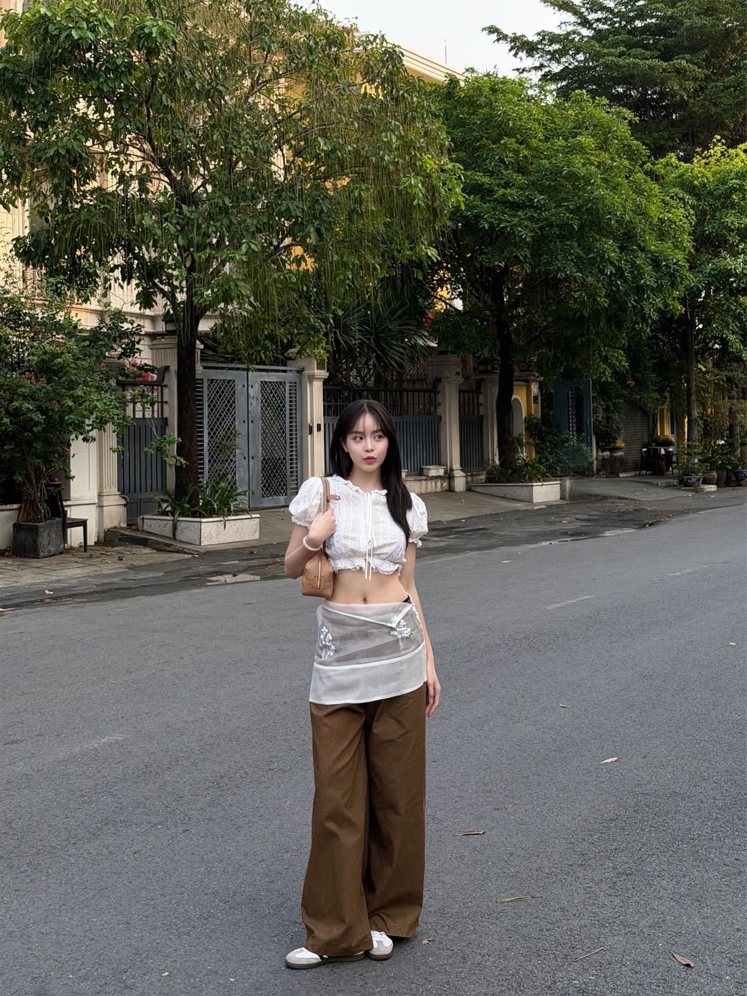 Thanh Thủy mix match chiếc áo crop-top trắng tay bồng với chiếc quần ống rộng màu nâu đất. Điểm nhấn là lớp khăn cột quần voan xuyên thấu khoác ngoài quần, tạo một tổng thể layer lạ mắt và thời thượng, mang đến diện mạo dạo phố vừa cá tính vừa high-fashion.