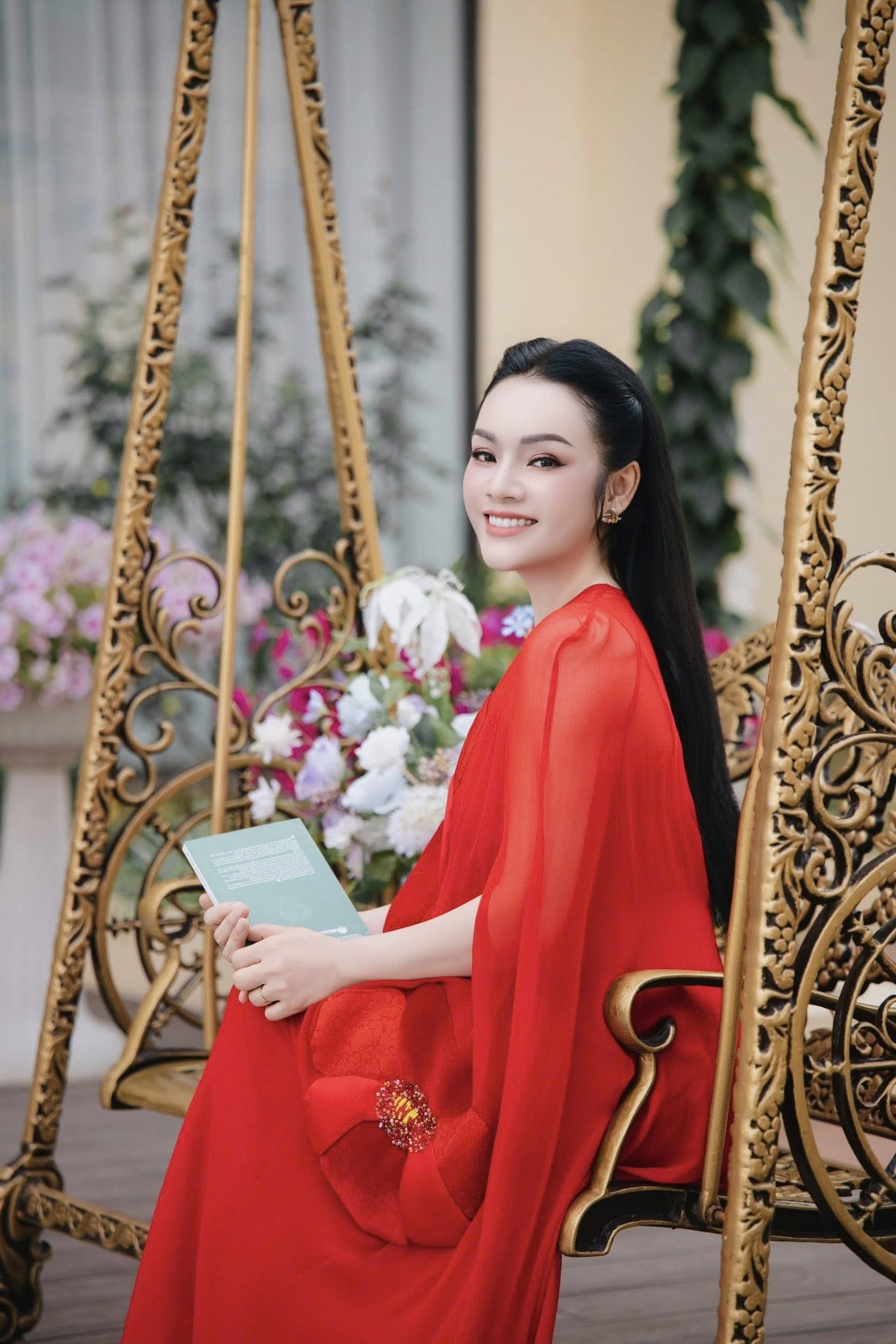 Trong khi đó, bộ váy đỏ lại tạo hiệu ứng hoàn toàn khác: nổi bật, đằm thắm và có phần quyền quý. Tông đỏ giúp làn da cô thêm sáng, kết hợp với thần thái điềm tĩnh tạo nên hình ảnh một người phụ nữ trưởng thành, từng trải nhưng vẫn giữ được nét mềm mại. Cách tạo dáng khi ngồi đọc sách giữa không gian nhiều hoa càng làm nổi bật vẻ đẹp tri thức, nhẹ nhàng mà không kém phần cuốn hút.