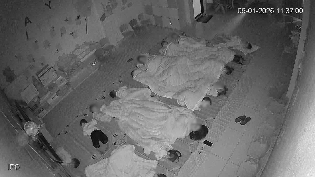 Trên người các con là những chiếc chăn to phủ chung. Ở góc gần cửa, hai bé trai nằm ngoài mép chăn, không được đắp gì.